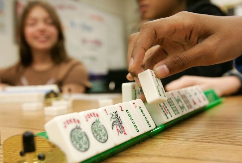 A child's hand places a Mahjong tile onto a green rack, with another child smiling in the background.