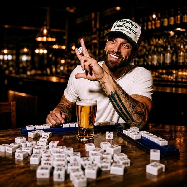 Smiling man with a beard and hat plays mahjong and drinks beer at a dimly lit bar table.